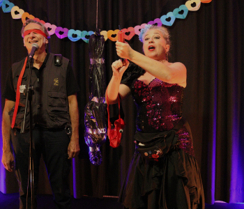 Mujer contando un monólogo en el escenario mientras un hombre del público está junto a ella con los ojos vendados, creando una atmósfera divertida y llena de sorpresa.